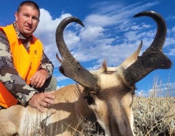 A hunter in an orange safety vest posing with a pronghorn antelope, set against a dramatic backdrop of fluffy white clouds and blue sky.